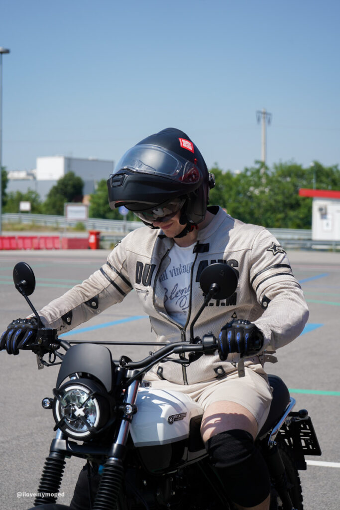 arge2rad-125ertesttag-arböfahrsicherheitszentrumwien-106 - I love my moped Motorcyclist wearing a helmet and gloves sits on a white and black bike in an outdoor lot under a clear blue sky.