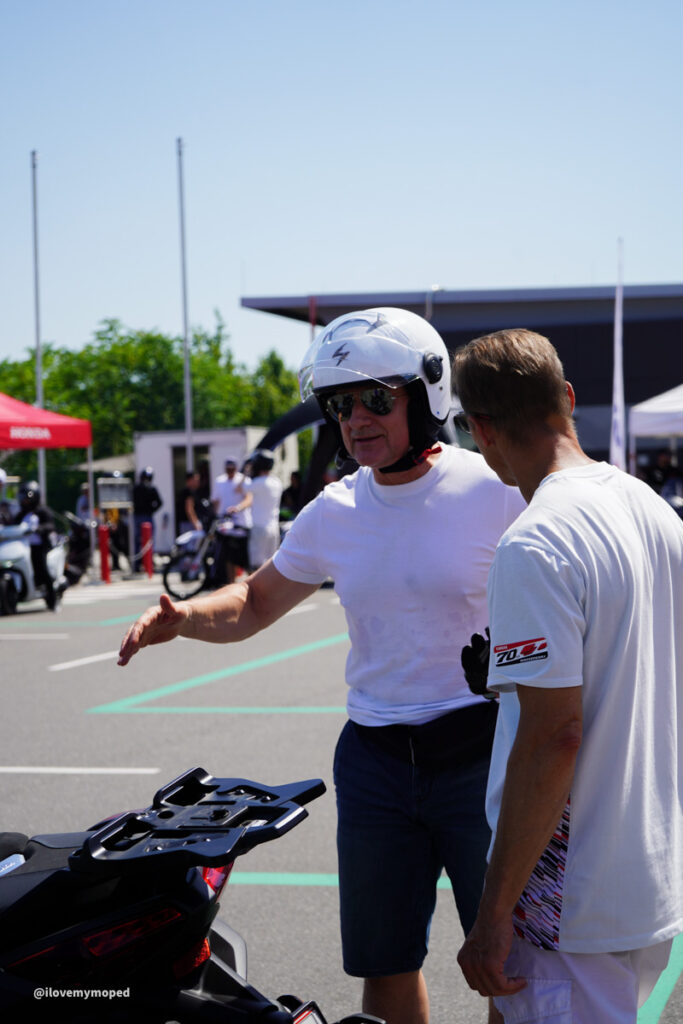 arge2rad-125ertesttag-arböfahrsicherheitszentrumwien-110 - I love my moped Two men talking beside a parked motorcycle at an outdoor event; one man wears a white helmet and sunglasses.