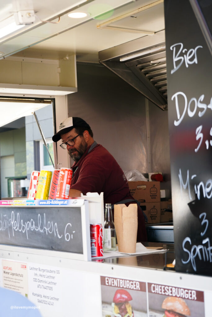 arge2rad-125ertesttag-arböfahrsicherheitszentrumwien-111 - I love my moped A bearded man in a baseball cap works behind a food truck counter with cans of drinks and a chalkboard menu on the side.